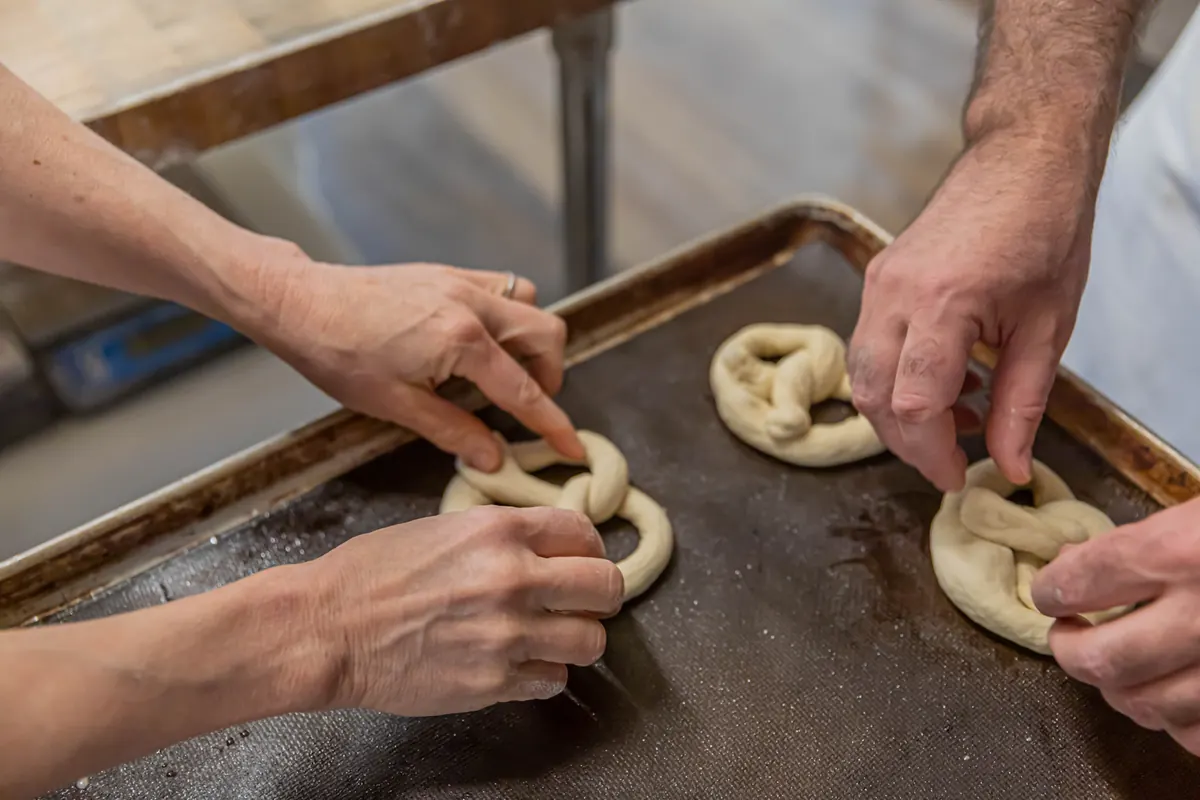 Two bakers twisting pretzel dough together on a baking sheet